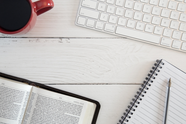 A Bible sits on table beside a cup of coffee, computer and notepad and pen