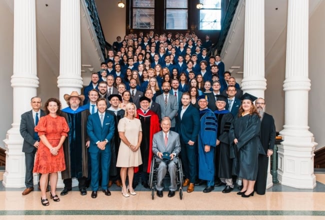 A group of University of Austin students in blue robes stand on stairs behind faculty and Texas governor Greg Abbott.