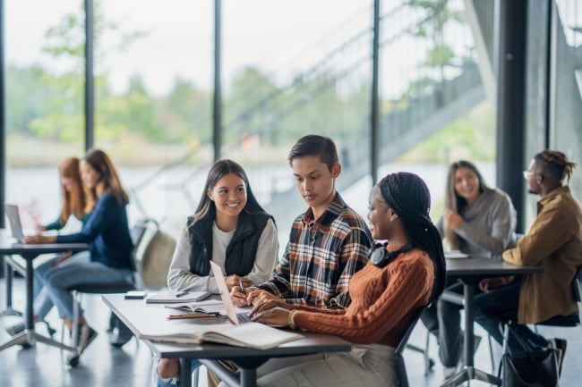 Three university students are seen sitting together in class as they work together on an assignment. They have a laptop open between them as they each give input for the assignment.