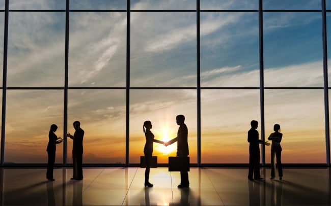 Three groups of two people stand before a large window looking out on the sun. They are shaking hands in greeting and talking together.