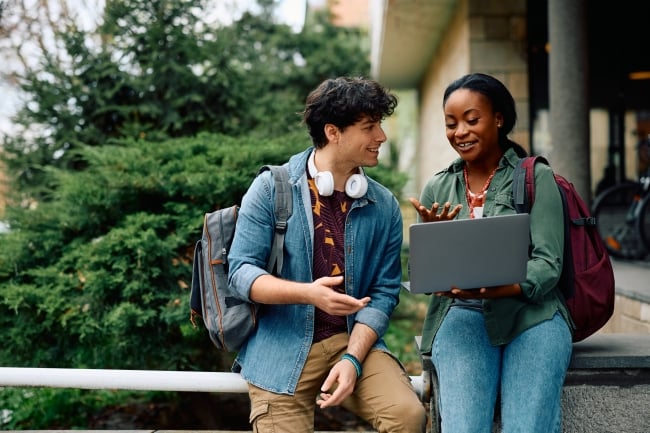 Two happy college students look at a laptop while wearing backpacks