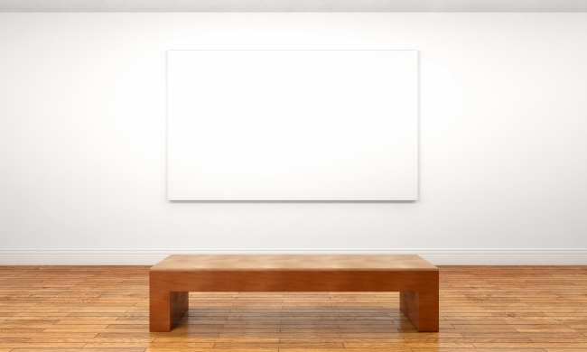 An empty wooden museum bench sits in front of a blank white canvas on a museum wall.