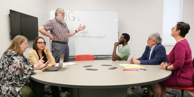 Mark Hoyert stands at a whiteboard with a group of five faculty members who are focused, considering his words