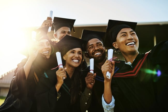 Five students pose in graduation caps and gowns holding diplomas and smiling.