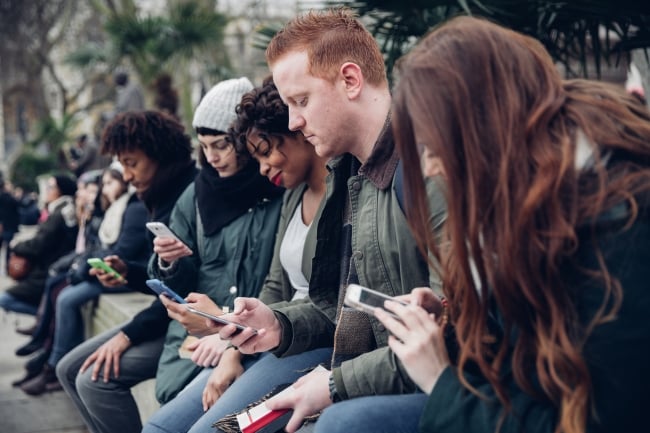 Group of students sitting outside on a ledge on a campus, all looking at their smartphones.