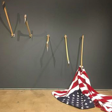 Art project shows a series of four empty flagpoles, from left to right, pointing up at a sharp angle, then at a less sharp angle, then drooping less than perpendicular. Fifth and final flagpole has an American flag attached, and it is pointing down so flag is dragging on a table.