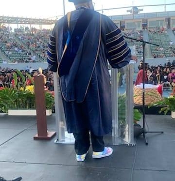 A man in academic regalia faces away from the camera, with a crowd of commencement guests in the background (in front of him).