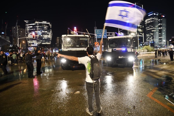 A protestor, holding an Israeli flag, seemingly blocking traffic, against a nighttime city backdrop.