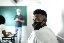 A young Black man wearing a black face mask over his nose and mouth looks over his shoulder. Behind him, a white professor wearing a mask is standing at a chalkboard.